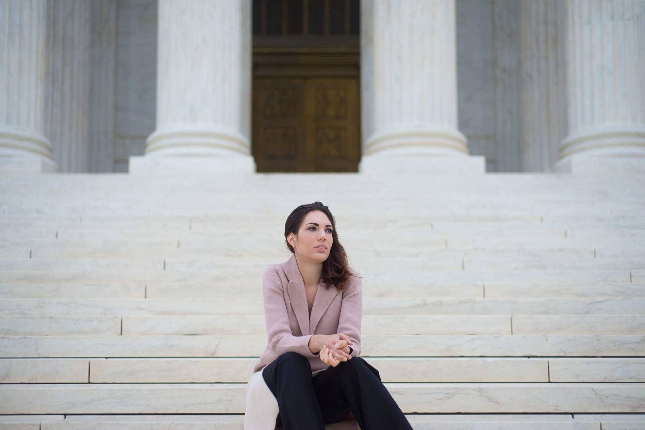 woman sitting on a flight of stairs wearing a beige jacket and denim pants