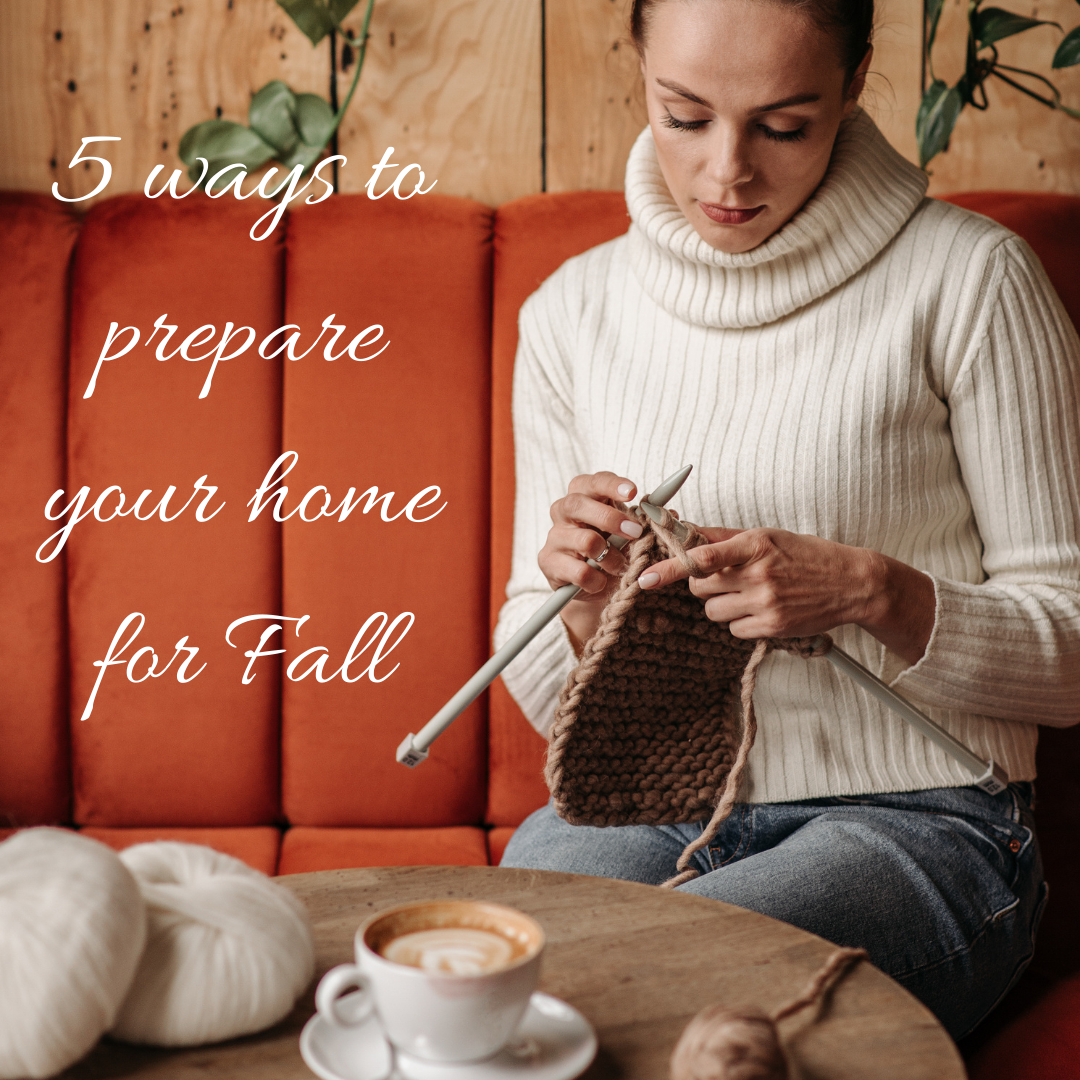 woman wearing a cowlneck sweater and denim pants. sitting an knitting on a chair