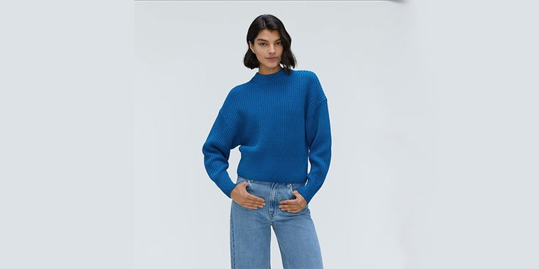 arafed woman in a blue sweater and jeans standing in front of a white wall